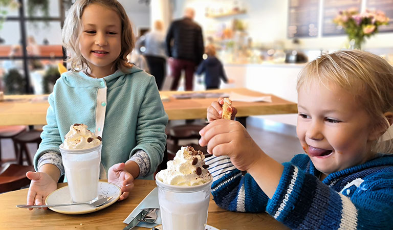Zwei Kinder in einem Cafe mit heißer Schokolade und großer Sahnehaube.