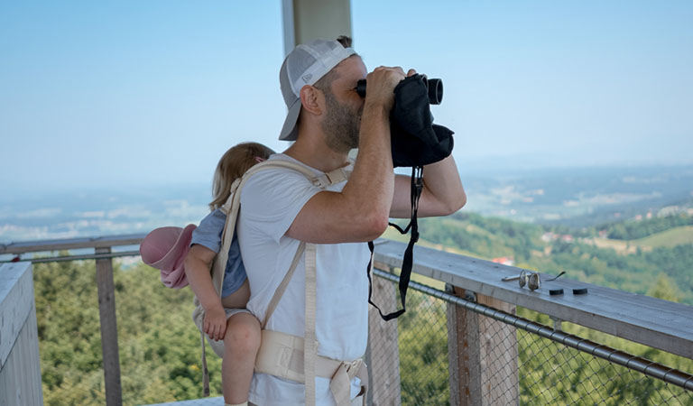 Vater mit Kind auf dem Rücken und Fernglas auf einem Hochstand in der >Steiermark in der Natur.