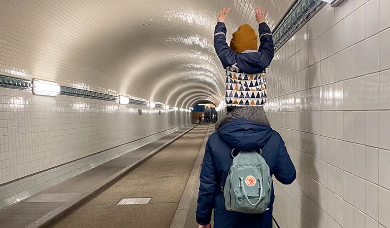 Kind auf den Schultern des Vaters im alten Elbtunnel mit Blick in die lange und weiß gekachelte Tunnelröhre.