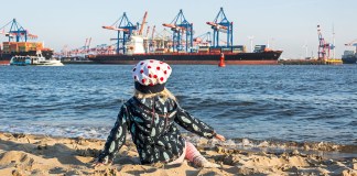Hamburg umsonst mit Kindern: 15 kostenlose Unternehmungen und Ausflugstipps für Familien Junges Kind sitzt am Elbstrand bei Oevelgönne und blickt auf ein Containerschiff am Ufer gegenüber.