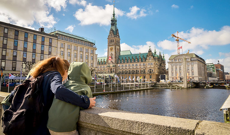 Mutter und Tochter stehen auf einer Brücke und schauen über einen Alsterfleet auf das Rathaus von Hamburg.